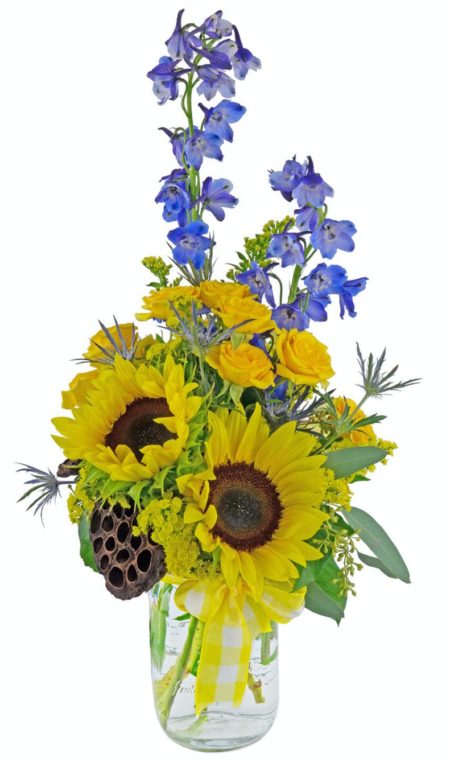 Blue and yellow flowers are arranged in a clear glass vase, and complimented with a yellow and white gingham ribbon. The flowers include blue delphinium and thistle, with yellow spray roses, sunflowers, and golden aster.