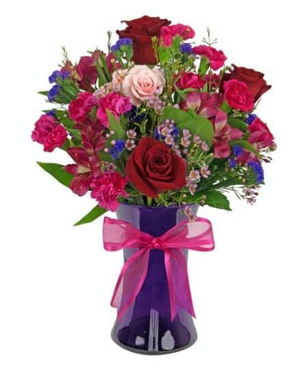 pretty purple vase contains red roses, pink alstroemeria, hot pink mini carnations, purple statice, pink spray roses, and waxflower.
