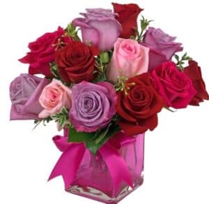 One dozen roses in red and shades of pink and lavender are artfully clustered in a pretty pink glass cube